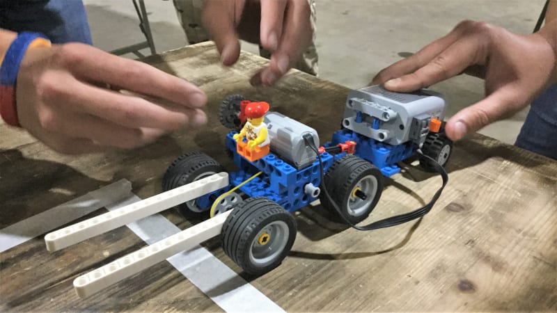 A LEGO vehicle that with various additions including a motor and trailer, two hands are working on the vehicle. 