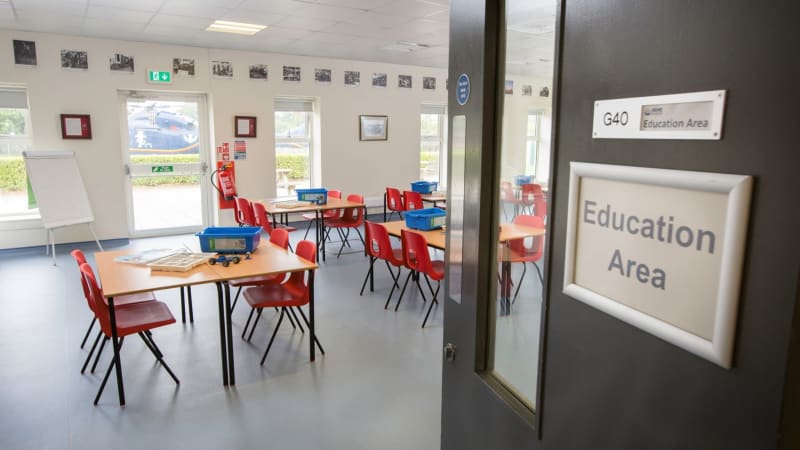A classroom taken through the doorway with door partially in the view, tables with 4 red chairs around each, blue boxes sitting on each.