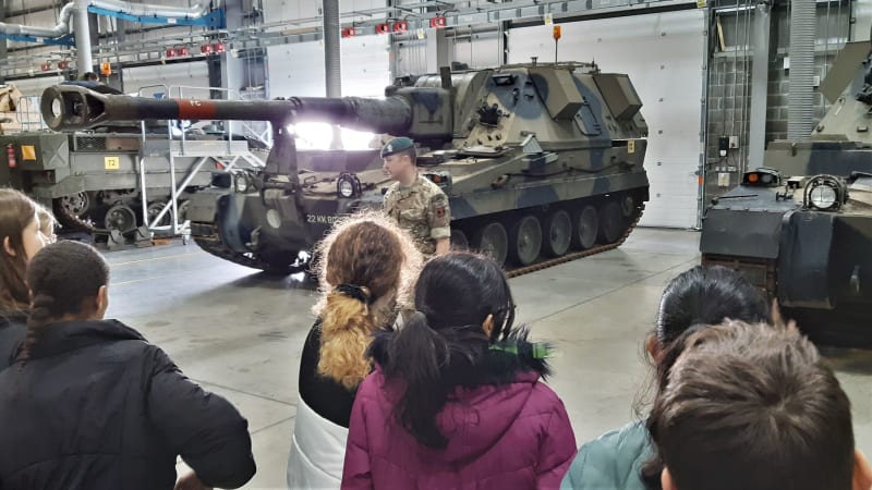 Children stood with their backs to the viewer, looking at an Army soldier in uniform, who is in front of a large tank inside a hangar. 
