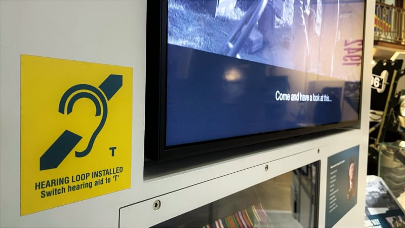 A yellow and blue hearing loop sticker on a wall next to a tv screen with subtitles showing at the bottom. 