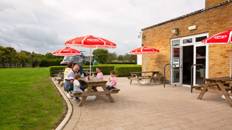 A family of two parents and three children sit at a picnic bench underneath a red umbrella. They are outside next to grass and hedges, and a brick cafe building is to the right.