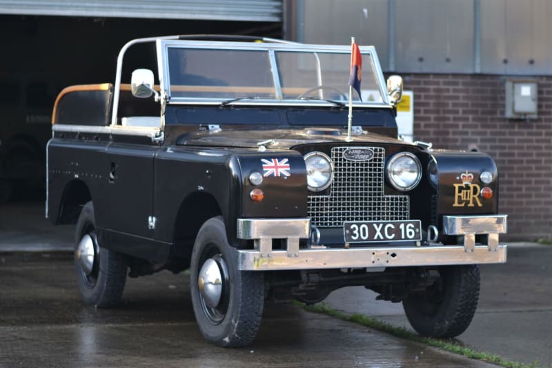 A black land rover with open cab from a diagonal perspective. British Union Jack and Queen Elizabeth IIs cypher are printed on the front, and a flag is on the bonnet.