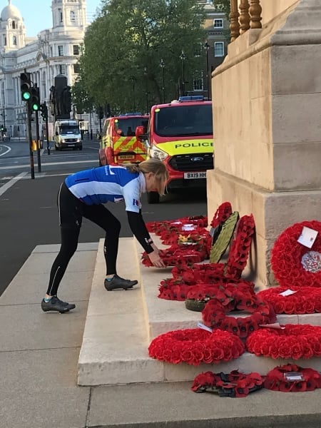 Woman in blue cycling jersey lays poppy wreath at base of white monument. Other wreaths surround. 