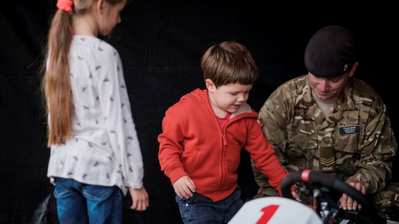A young boy in red hoodie touches the wheel of a go-kart. A man in military camouflage bends down beside him. A young girl in a white top looks on with back to camera.