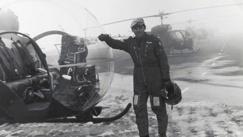 Black and white photo of a pilot in uniform leaning against a helicopter. He holds a helmet in his left hand. More helicopters out of focus on runway behind.