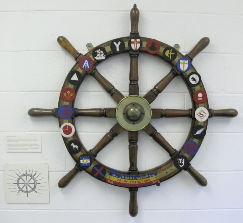 Wooden wheel with eight spokes and twenty badges attached to the front around the edge of the wheel. Mounted on a white brick wall.