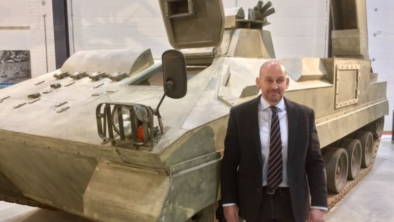 Major Henderson in jacket and tie stands in front of a tank inside the museum.