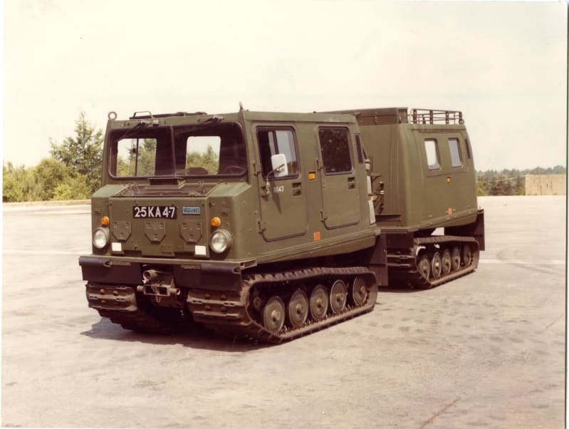 A tracked vehicle with tracked trailer, on snowy ground, trees in the background.