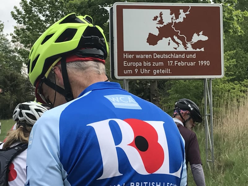 Cyclist in blue jersey and yellow helmet stands with back to camera. Two other cyclists in background. Brown sign with map of Europe and German writing in top right corner of photo.