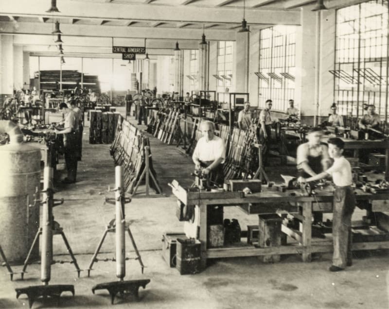 Black and white photo of men working at stations with bits of material in a warehouse. 