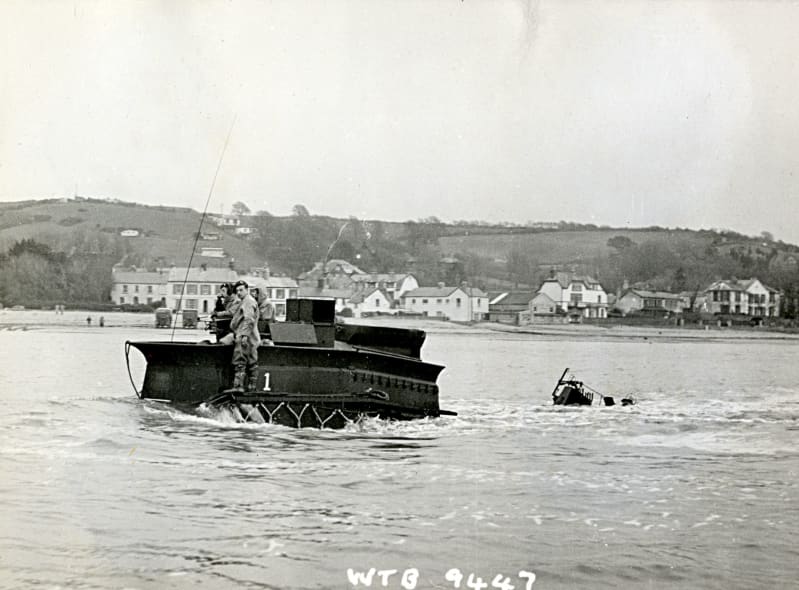 A vehicle is half submerged in water, soldiers stand on top of it. Houses and hills in background.