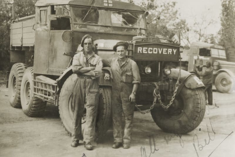 Sepia photograph of two men in overalls and berets stood in front of a large heavy vehicle with " recovery " on front. 