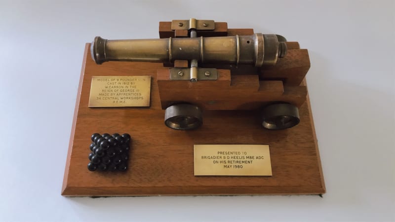 Aerial view of a miniature canon and canon balls made from wood and metal. They are on a flat wooden base with two metal rectangles with inscriptions.