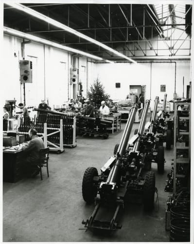 Black and white photo of men working at stations in a workshop. Large guns on wheels line the side of the room. 