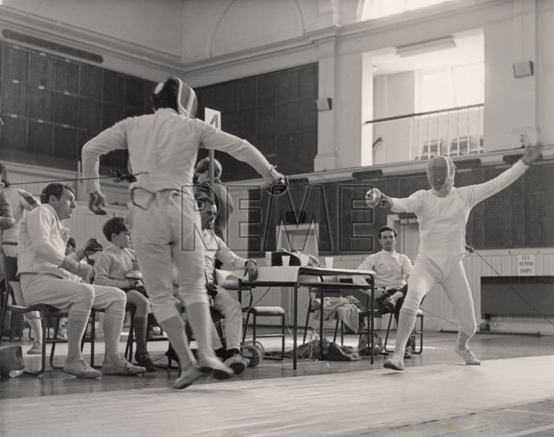 Two people in fencing kit are in the middle of a fencing match. Others wear fencing kit and sit at tables in the background watching on.