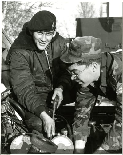Two soldiers working on an engine, one looks at the camera.