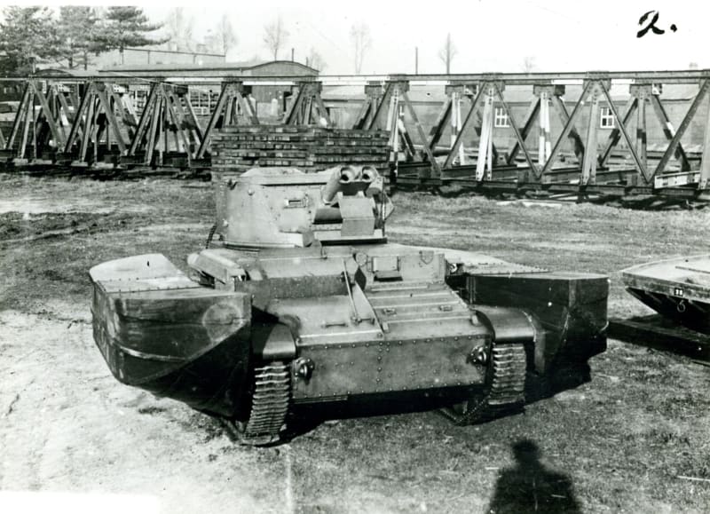 Black and white photograph of a tank with large side attachments, buildings and framework in background.