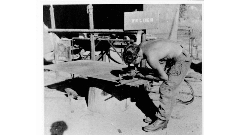Black and white photograph shows a man, topless, leaning over a bench cutting material, a sign in the background reads 