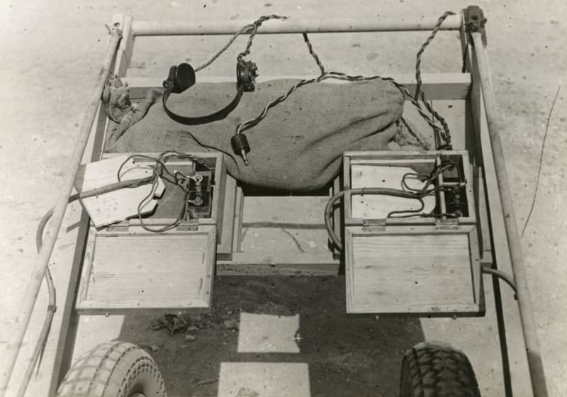 Image shows a metal frame on wheels, with two wooden boxes on top, wires and headphones. 
