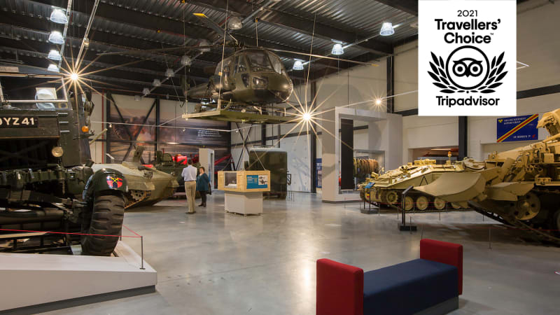 Large room with tanks, trucks and a suspended helicopter on display, a red and blue bench in the foreground. Black and white logo in the top right corner reads "2021 Travellers' Choice". 