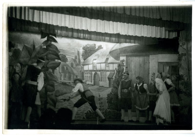 Black and white photo of six actors on a small stage with village scenery painted behind. A large prop beanstalk is on the stage. 