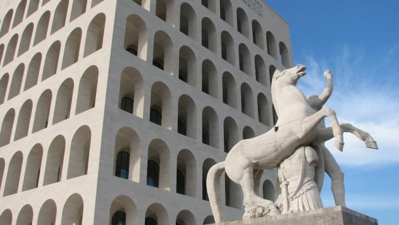 A white marble cubed colosseum building in the background with many windows. A white marble statue of a horse rearing and a soldier in Roman uniform in the foreground.