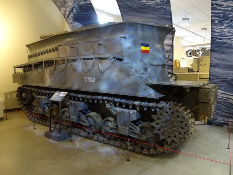 Image of an armoured recovery vehicle with tracked wheels, grey colour with square of blue yellow and red horizontal stripes on the front.
