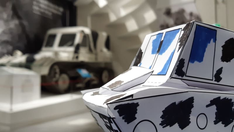 Paper model of tracked vehicle in the foreground with black and white tracked vehicle blurred in the background.
