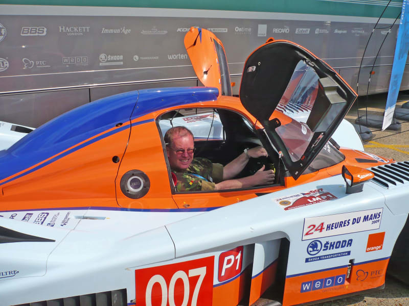 A REME officer in uniform sat inside a racing car (side view) looking at the camera, with orange white and blue livery, small doors opened outwards and upwards.