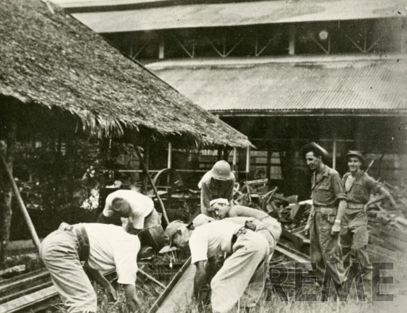 Black and white photo outside some huts, one with straw roof. Men dressed in white are doing some form of manual labour while 2 soldiers look on.