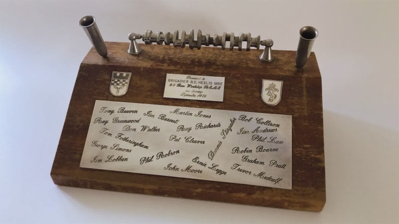 Dark brown wooden box with a slanted side at the front. A metal crank shaft with two pen holders either side on top. Two coats of arms and a dedication in metal at the top of the slanted side. Large metal rectangle with various names engraved underneath.