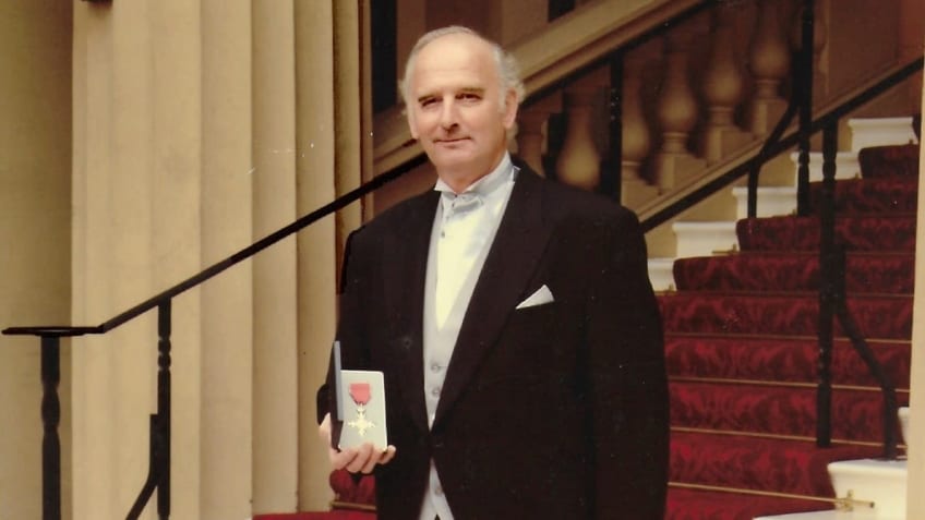 Sergeant Jones in black suit stood on red carpteed stairs and holding and MBE award.