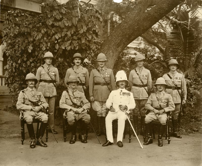 Black and white photograph of two rows of Army Officers in uniform with Pith helmets (half sitting half standing). Most have moustaches. 