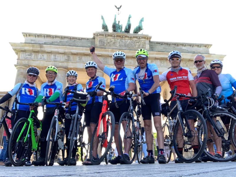 Row of eight cyclists in blue or red jerseys stand with bikes in front of German building. Man in middle raises one arm in the air. All look at camera.