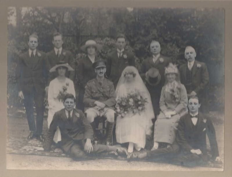 Black and white photo of a group of people in suits and dresses, in centre bride in white wedding dress and groom in army uniform.