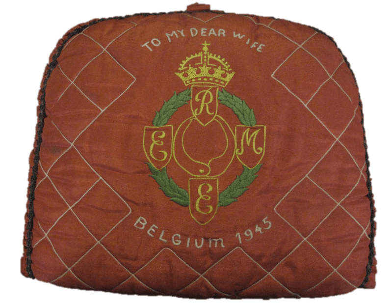 Dark red tea cosy with white quilting and first pattern REME badge embroidered in yellow and green thread. White embroidery above and below the badge reads "To my dear wife Belgium 1945" .
