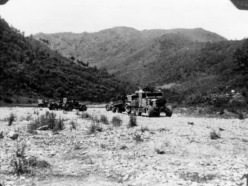Vehicle driving along a dry river bed with another vehicle in tow, with hills flanking either side. 