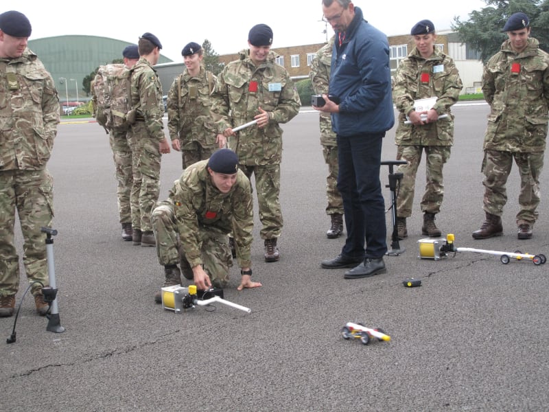 Soldiers standing around and one crouching next to an air pump, where a toy vehicle is moving away from.