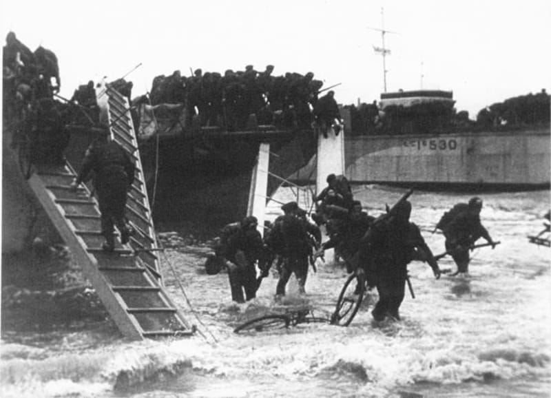 Black and white photo of soldiers disembarking landing craft on a beach shore, some have disembarked with bicycles.