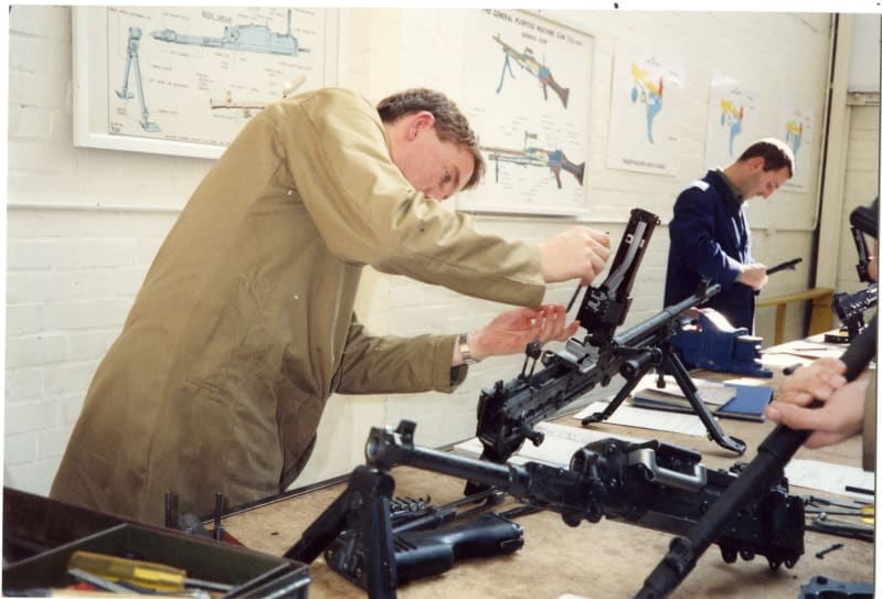 Man in a brown jumpsuit works on a machine gun at a desk, which has other parts on it. A man in the background in a blue jumpsuit works on another gun. The hands of someone holding a gun are seen in the foreground. Posters of the inner workings of guns and machines on wall in background.