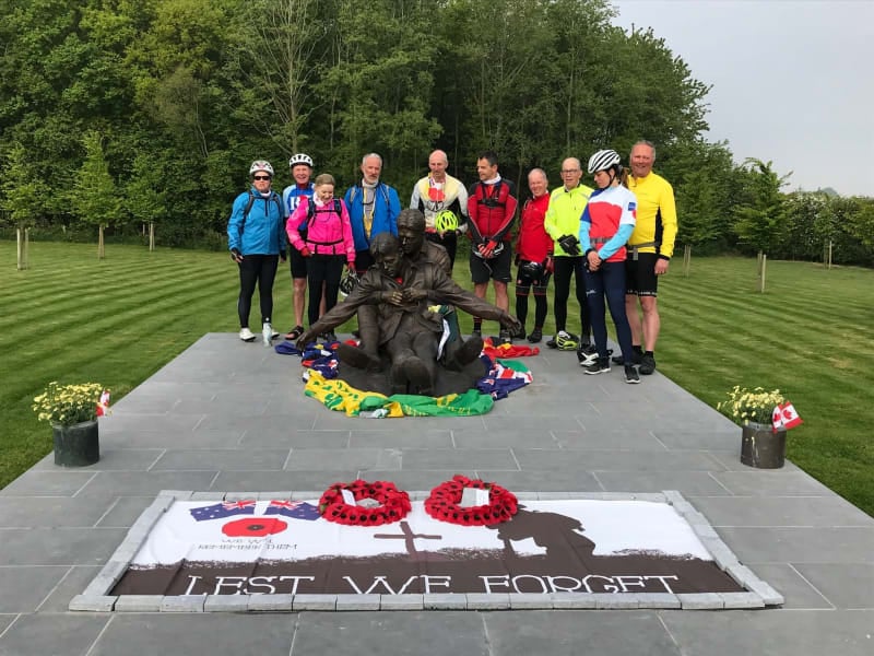 Group of cyclists in a field gather in line behind a statue of a soldier carrying another. In front, two poppy wreaths and a memorial on the floor reads " Lest We Forget ". 