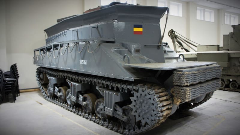 Large grey tracked vehicle with a small Belgian flag painted on the front. Parked next to another tracked vehicle in a room. 