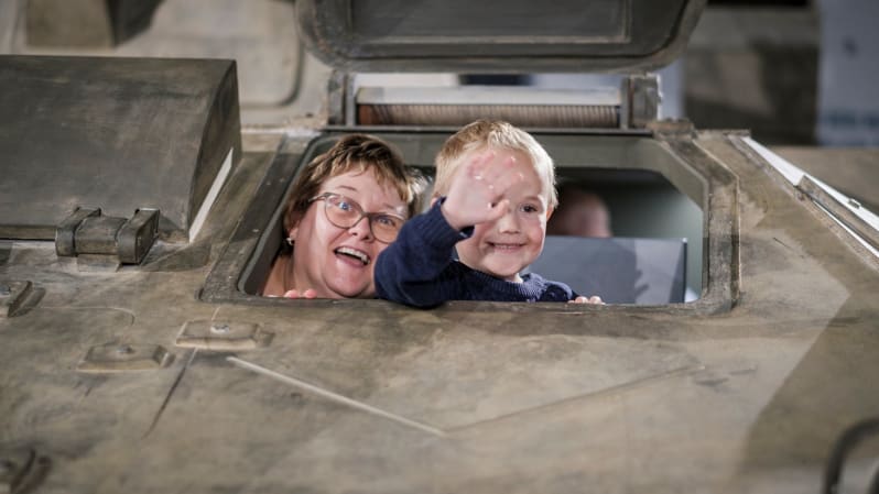 Close up of a woman and young boy poking their heads out of a tank. Both smile at camera and boy waves.