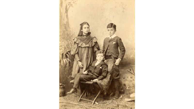 Black and white photograph of three children, two standing and one on a chair in front.