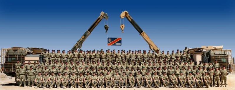Large group of men in army uniform sit or stand in rows outside. All face camera. Two cranes behind hold up blue flag with red stripe. 