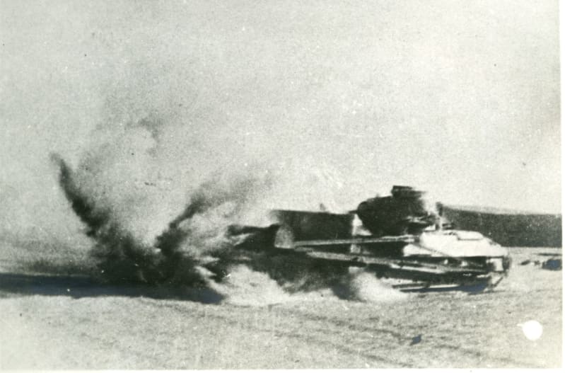 Black and white image. Side view action shot of a tank in the desert, visible is a large cloud of dust surrounding the tank which is slightly blurred from movement.