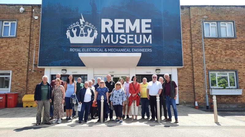 A group of people stand posing for a photo outside of a building with large blue sign reading " REME Museum " in white text. 