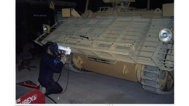 Image shows a metalsmith kneeling in front of a tracked armoured vehicle, there is a bright light where they are working.