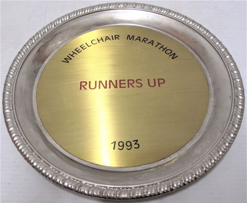 Metal circular plate with silver patterned edge and gold coloured in the centre. Reads " wheelchair marathon runners up 1993 " 