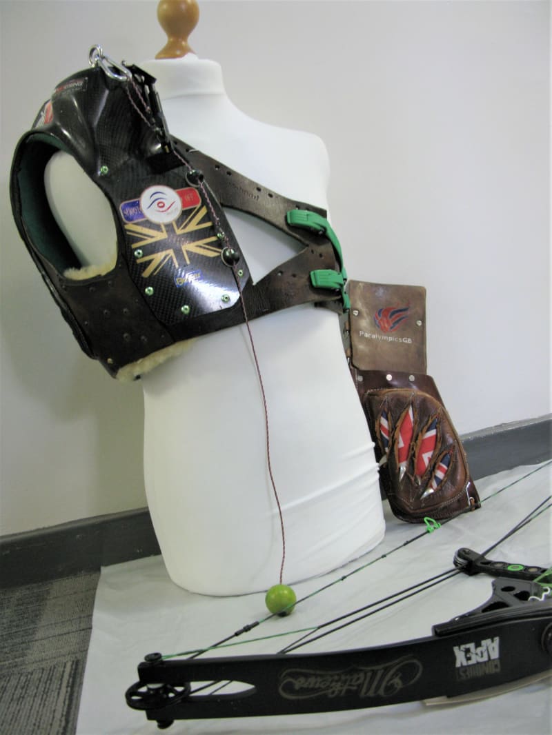 Photo of archery equipment, including a mannequin with specialist para-athlete brace on the shoulder/chest, a quiver to the side and bow laying in front.